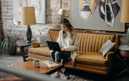 Eine junge Frau sitzt auf einem senffarbenen Sofa und schaut auf ihren Laptop