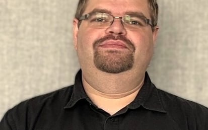 Eine Person steht vor einem neutralen, strukturierten grauen Hintergrund. Die Person trägt ein kurzärmliges, schwarzes Hemd mit Knopfleiste und eine dunkle Smartwatch am linken Handgelenk. Die Arme sind vor dem Körper locker verschränkt. Die Person hat kurzes, braunes Haar, trägt eine Brille mit schmalem Metallrahmen und einen gepflegten Kinnbart. Der Gesichtsausdruck wirkt ruhig und freundlich. Es sind keine marken- oder barrierebezogenen Elemente im Bild sichtbar.