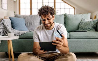Ein junger Mann sitzt auf einem Teppich in seinem Wohnzimmer, schaut auf sein Tablet und hält eine Kreditkarte in einer Hand