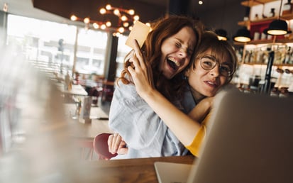 Zwei junge Mädels umarmen sich lächelnd in einem Café an einem Tisch und freuen sich über ihren erfolgreichen Black Friday Kauf