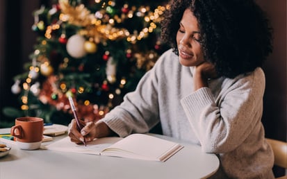 Eine Frau sitzt entspannt an einem Tisch vor einem festlich geschmückten Weihnachtsbaum, schreibt in ein Notizbuch und trinkt aus einer Tasse.
