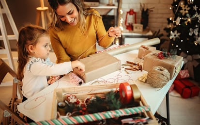 Eine Mutter und ihre Tochter verpacken gemeinsam Weihnachtsgeschenke während im Hintergrund ein geschmückter Weihnachtsbaum für festliche Stimmung sorgt.